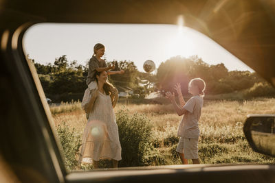 Mother and sons playing with soccer ball standing in meadow seen through car window