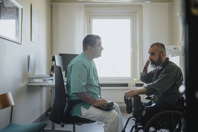 Side view of mature male doctor discussing with patient sitting in wheelchair at clinic