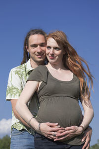 Smiling young man embracing pregnant woman while standing against blue sky