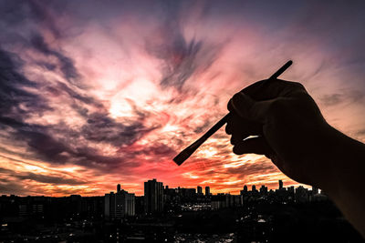 Close-up of silhouette hand holding paintbrush against city during sunset