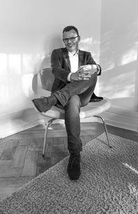 Full length portrait of smiling businessman sitting on chair against wall at home