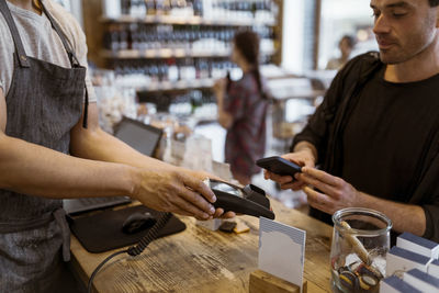 Midsection of male owner giving credit card machine to customer doing online payment at store