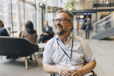 Smiling businessman with disability holding coffee cup looking away while sitting on wheelchair at convention center