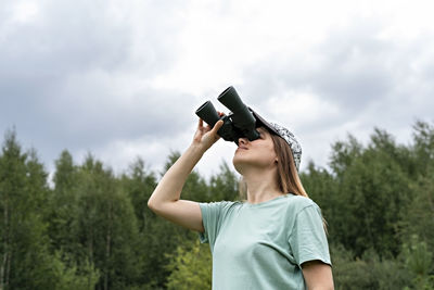 Young blonde woman bird watcher in cap  looking through binoculars cloudy sky in forest ornithology