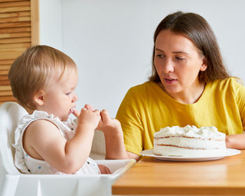 Baby looking at creamy cake with interest while sucking finger. mother helping daughter eat pie