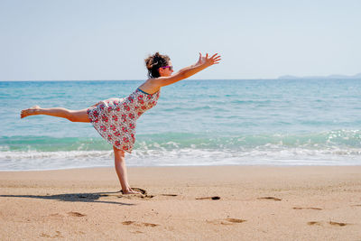 Woman with arms raised exercising while standing on one leg at beach against sky