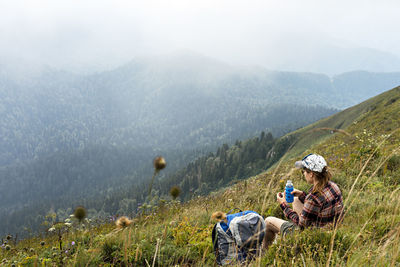 Young woman sitting on meadow in mountain valley next to backpack drinking water  on hike landscape 