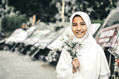Portrait of smiling girl wearing white hijab holding flowers on street