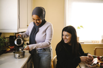 Smiling young woman looking at friend pouring tea standing at kitchen counter