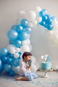 Baby boy in blue pants and shirt standing on the floor next to the cake in blue and white balloons