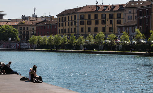 People relaxing by lake against buildings in city