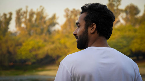 Side view of young man looking away against trees