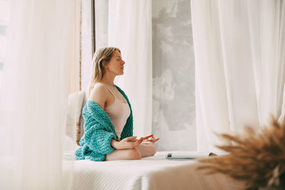 A young woman in pyjamas is sitting on a bed in a lotus position with her fingers folded in a mudra 