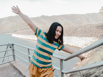 Portrait of smiling young woman with arms outstretched standing by railing