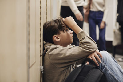Depressed teenage boy sitting by locker against school corridor