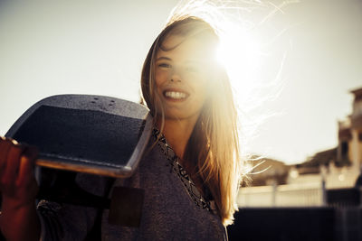 Portrait of smiling young woman holding skateboard during sunny day