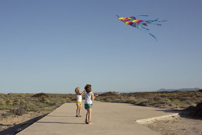 Full length of delighted little children in shorts walking on road and smiling while launching colorful kites against cloudless blue sky on sunny summer day