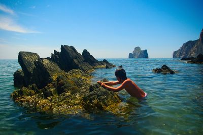 Side view of girl on rock in sea against blue sky
