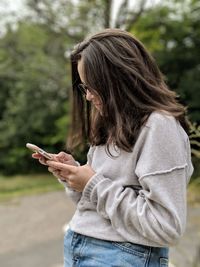 Side view of woman using phone while standing against trees