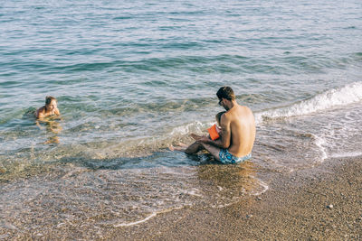 Rear view of shirtless man sitting at beach