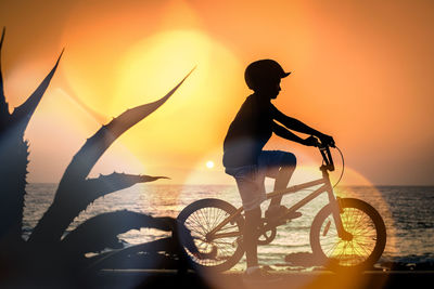 Silhouette of a young male on a bicycle with sea and orange sunset in background boy enjoying summer