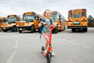 Funny smiling happy caucasian girl kid child with backpack riding scooter on school yard 