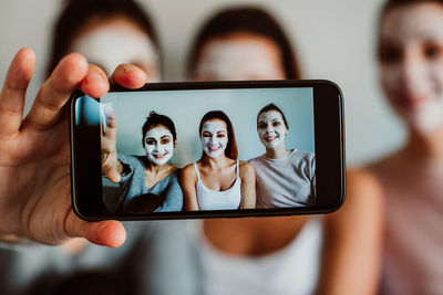 Female friends with facial masks taking selfie against wall at home