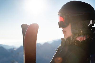 Portrait of sportswoman in helmet and mask with skis in hand, looking away, enjoying sunny frosty