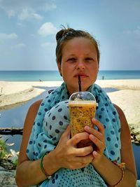 Portrait of young woman holding ice cream at beach against sky