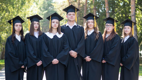 Seven graduates in robes stand in a row outdoors