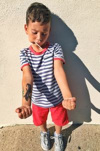 Full length of cute boy showing tattoo while eating lollipop against wall