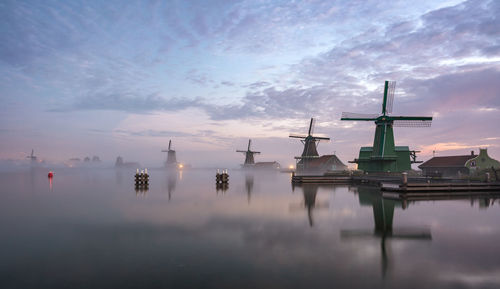 Traditional windmill at lakeshore against sky during sunset