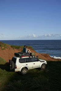 Mid adult couple sitting on 4x4 roof while looking at sea against blue sky