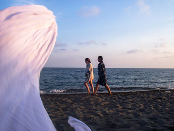 Friends with towels walking at beach against sky during sunset