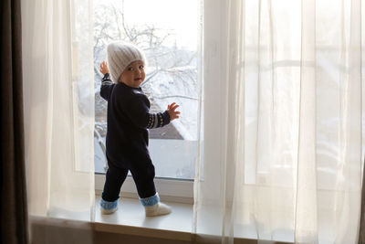 Child in a warm blue jumpsuit is on window in winter white knit hat