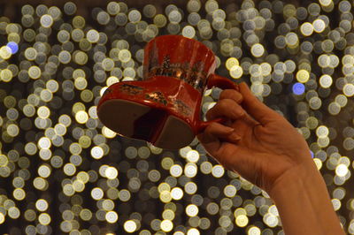 Cropped hand of woman holding shoe shaped mug against lights