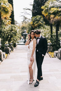 Happy lovers the bride and groom in wedding outfits walk among plants and palm trees in the old park