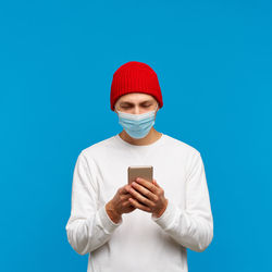 Man wearing mask using smart phone standing against blue background