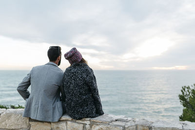 Rear view of couple sitting on retaining wall by sea