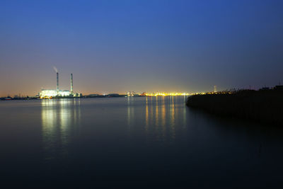 Illuminated factory by river against sky at sunset