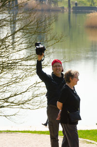Portrait of man holding camera standing with wife by lake
