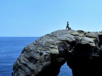 Man standing on rock by sea against clear blue sky