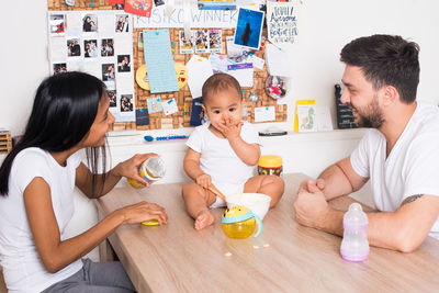 Parents looking at baby having food while sitting on table at home