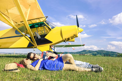 Young couple lying on grass by airplane on field against sky