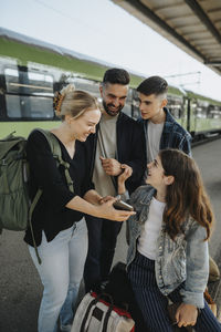 Happy woman showing smart phone to family while waiting for train at railroad station