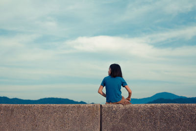 Rear view of girl sitting on retaining wall against sky