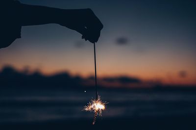 Silhouette hand holding fireworks against sky during sunset