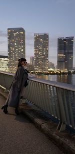 Side view of mid adult woman standing on footbridge over river against sky in city during sunset