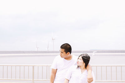 Young couple looking away while standing against railing and sea