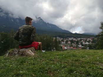 Rear view of man sitting on landscape against cloudy sky